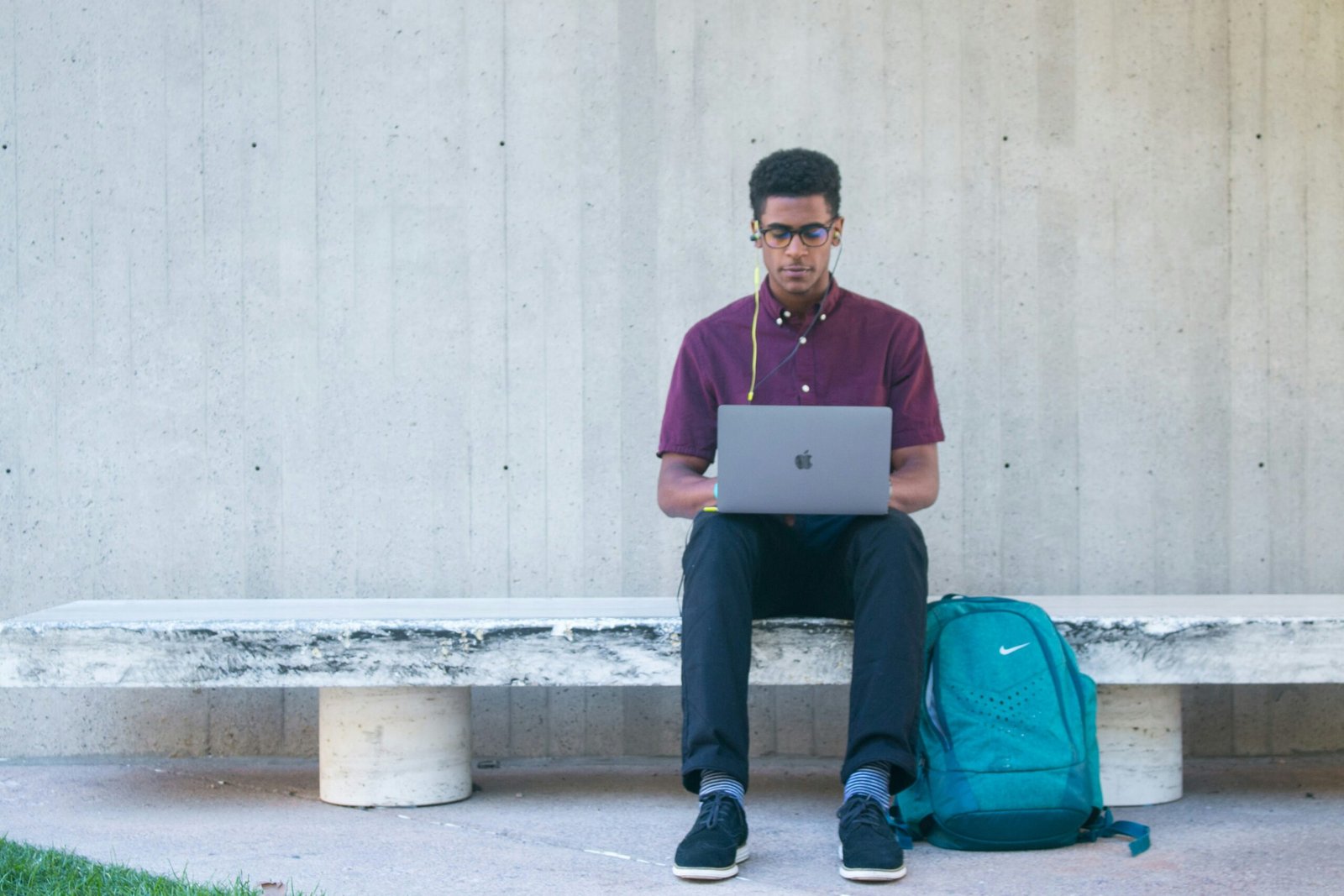 A South Asian man sitting on a bench with earphones plugged Into a MacBook.
